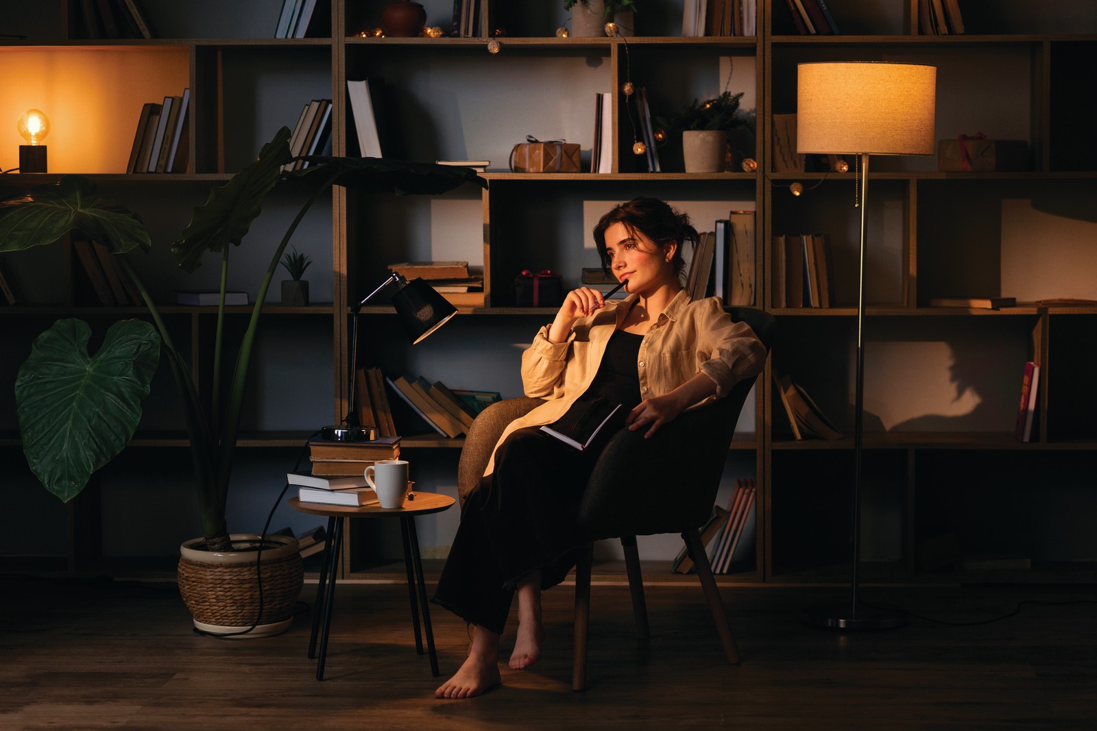 Moody by Design – Into the Dark A woman relaxes in an armchair in a dark, cocooning library lit by warm lamps