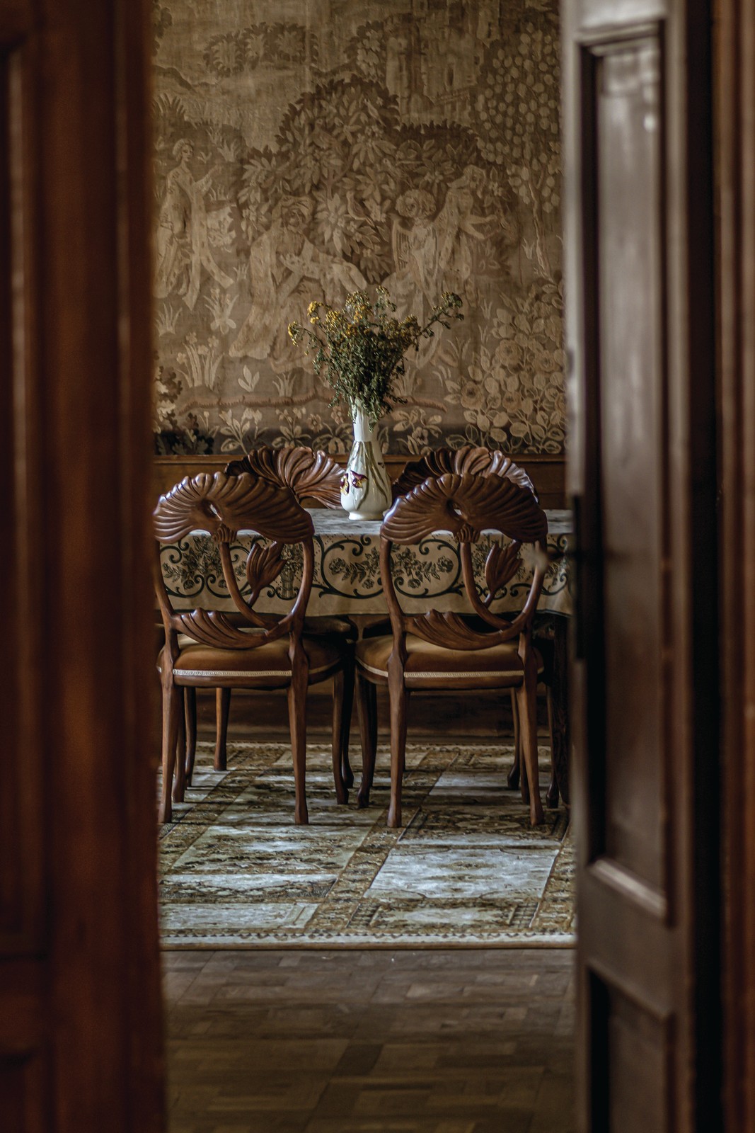 Old-World Revival – Heritage Reimagined View through a doorway into an old-world dining room with carved chairs, a patterned rug and textured walls
